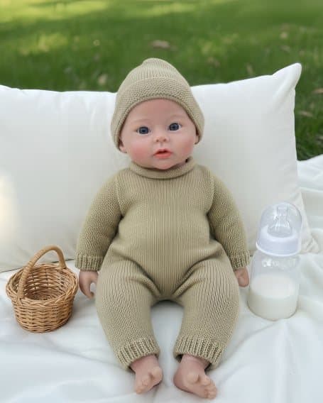 Reborn doll in a beige knit hat and knit jumpsuit, sitting against a white pillow beside a small basket and a milk bottle.