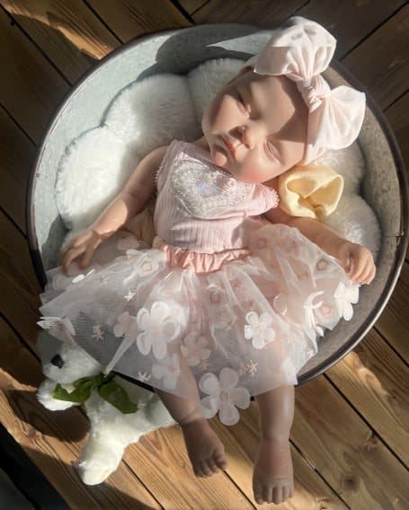 Sleeping reborn doll in a pink tulle dress and bow headband, curled in a round bowl on a wooden deck.
